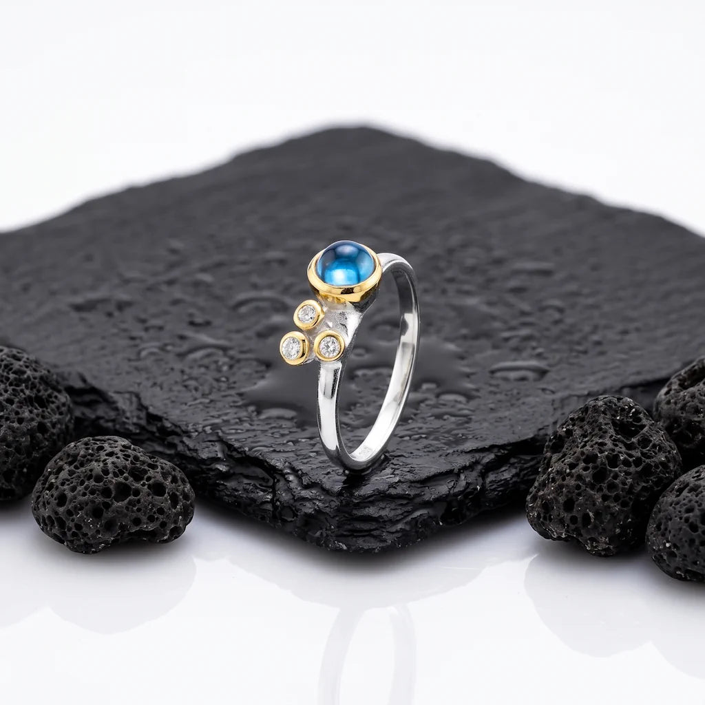 ARTEMIS Aura Ring – Exklusiver Bicolor Ring mit irisierendem Spektrolith und blauem Topas aus 925 Sterling Silber präsentiert auf schwarzem Lavastein von René Rose Jewelry
