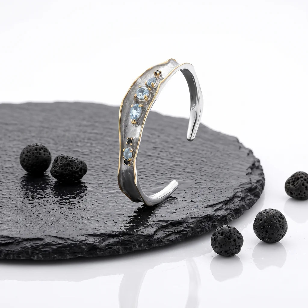 ARTEMIS WAVE – Offener Bicolor Armreif im Wellen Design mit blauem Topas aus 925 Sterling Silber präsentiert auf schwarzem Lavastein von René Rose Jewelry