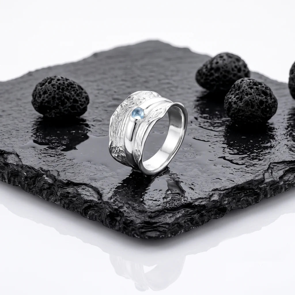FENNA GLACIER – Breiter Statement Ring mit blauem Topas Edelstein aus 925 Sterling Silber präsentiert auf schwarzem Lavastein von René Rose Jewelry
