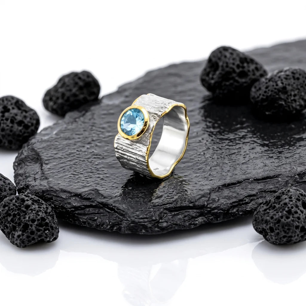 ISHANI GLACIER – Breiter Bicolor Statement Ring mit blauem Topas Edelstein aus 925 Sterling Silber präsentiert auf schwarzem Lavastein von René Rose Jewelry