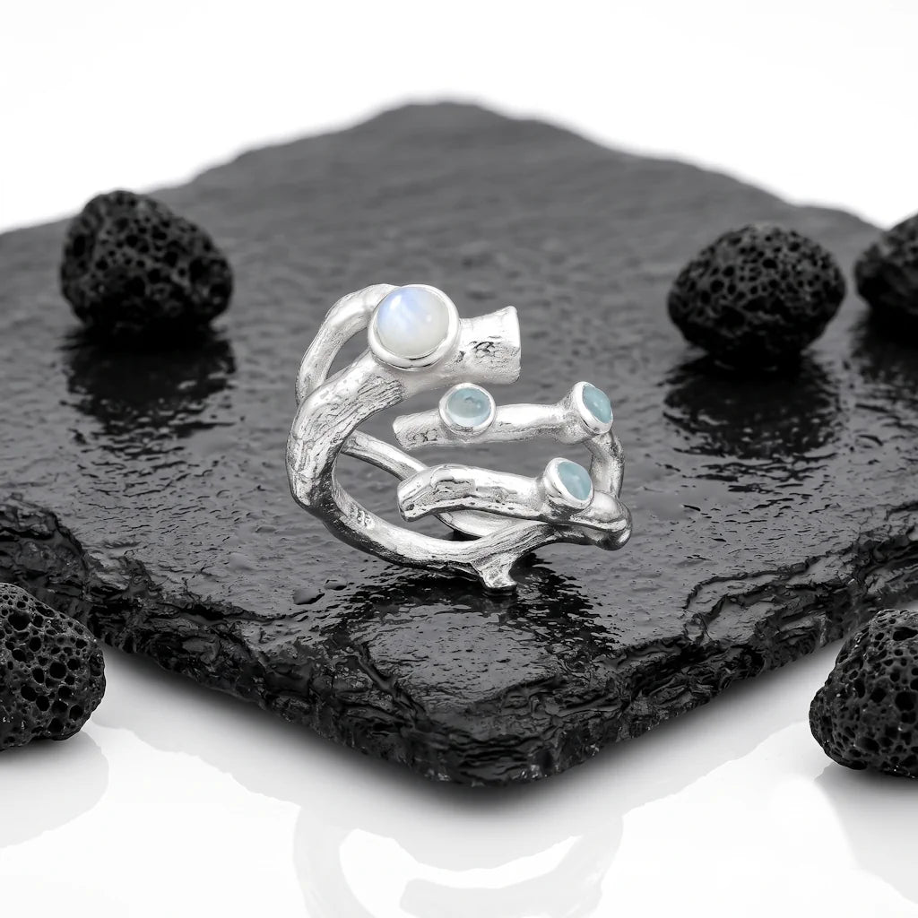 SUNNIVA ARKTIS – Breiter Statement Ring mit Regenbogenmondstein und blauem Topas aus 925 Sterling Silber präsentiert auf schwarzem Lavastein von René Rose Jewelry