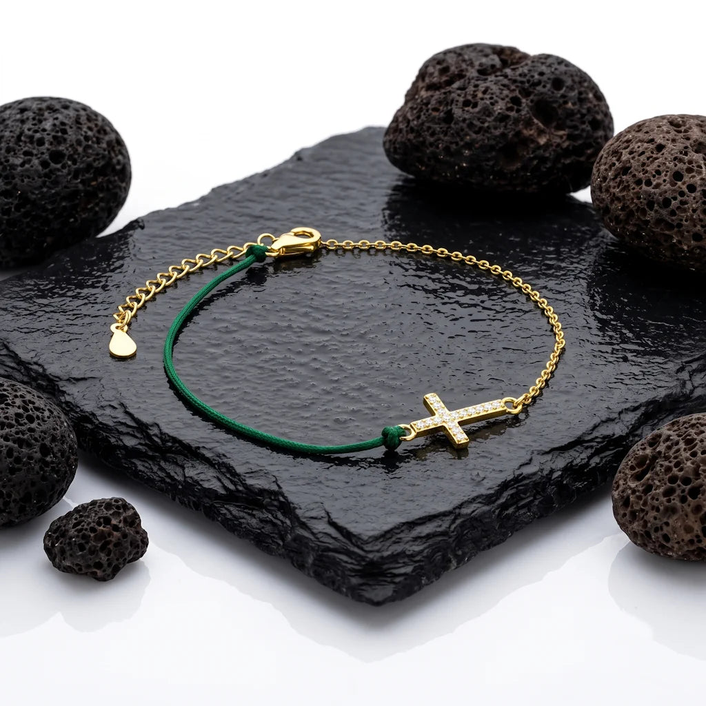 René Rose Jewelry SERAPHINE luxuriöses Designer-Armband aus 925 Sterling Silber mit 24 Karat Gelbgold-Vergoldung, weißen Zirkonia, Kreuz-Anhänger und dunkelgrüner Seidenkordel, stilvoll auf Lavastein und Schieferplatten präsentiert.